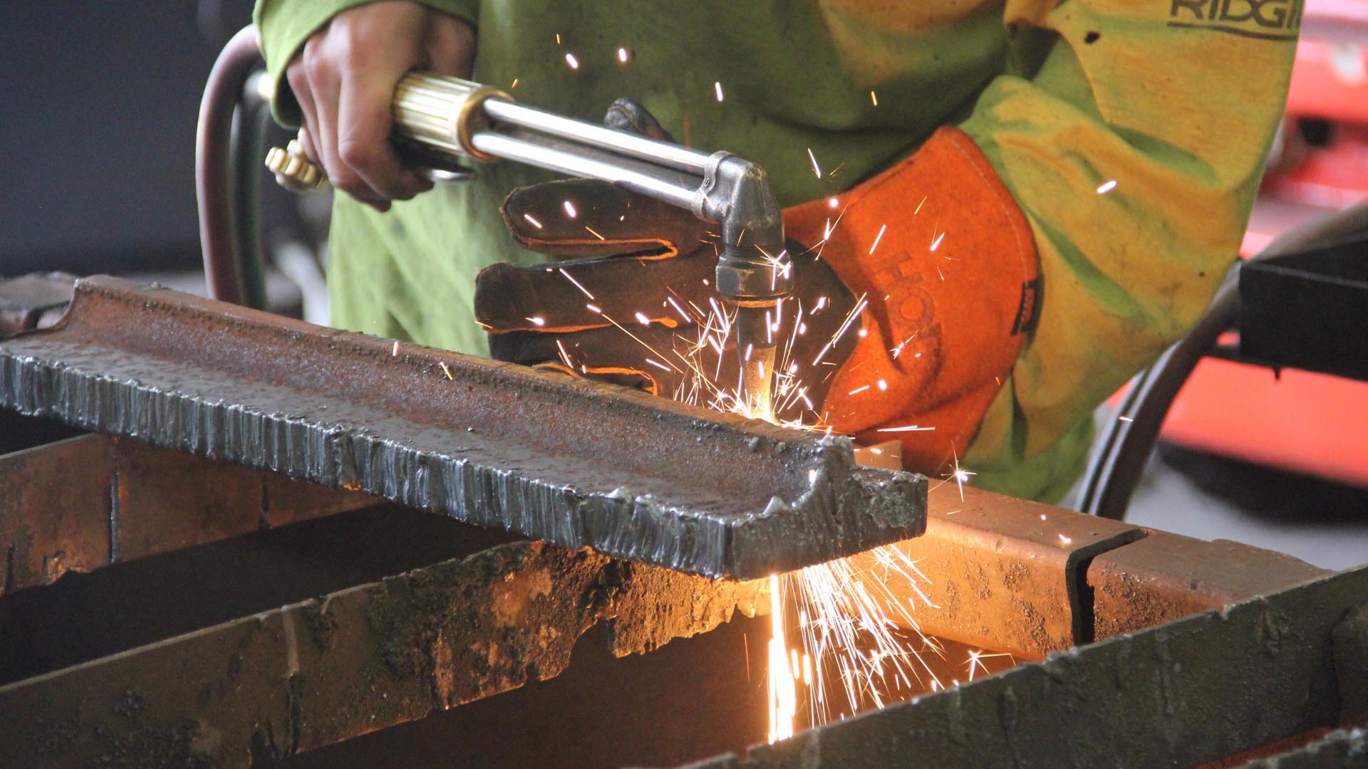 Worker wearing protective gloves using a torch to cut or weld a piece of metal, sparks flying in a manufacturing setting