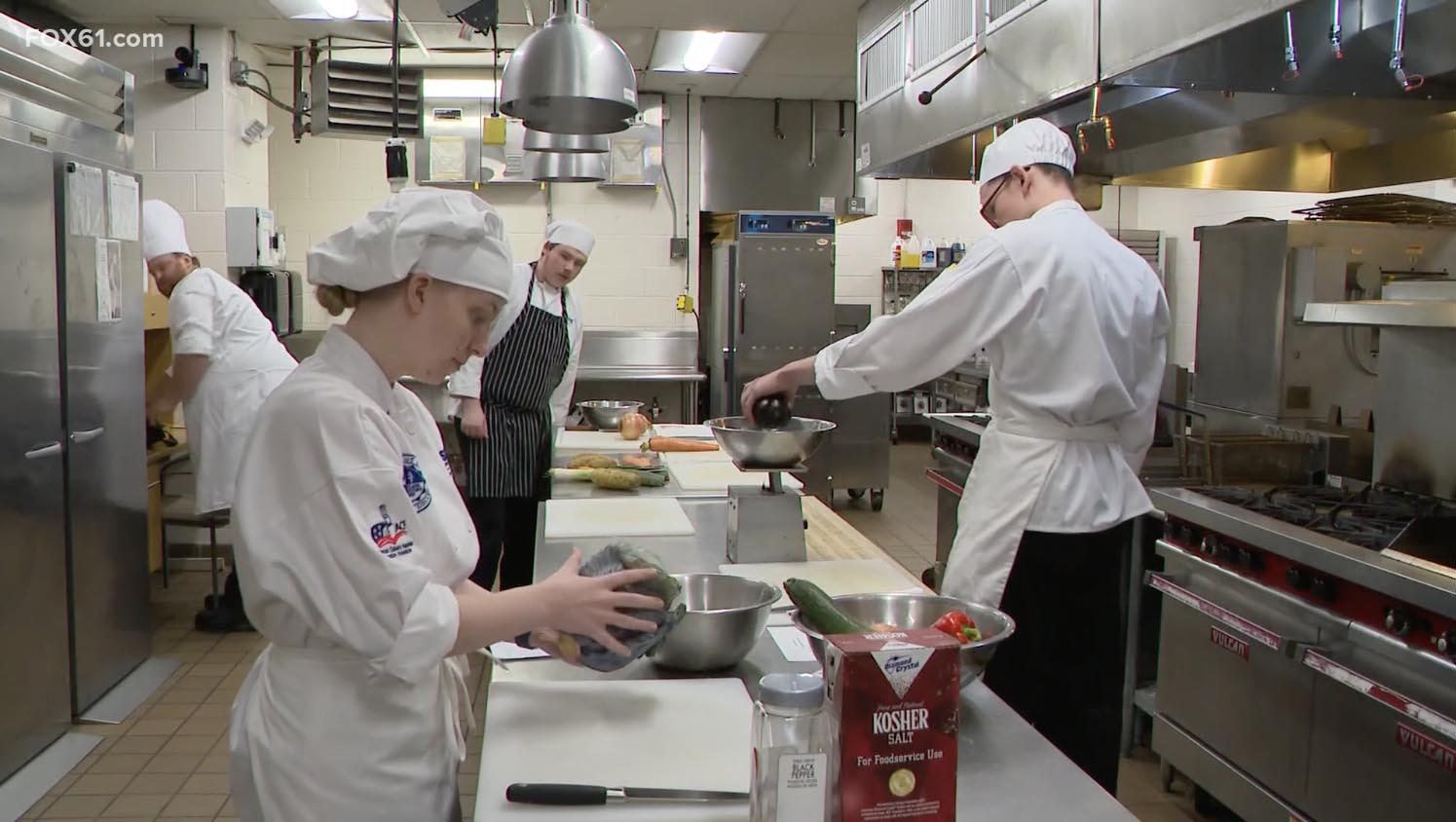 CT State culinary students in uniform chop vegetables in a professional kitchen during a cooking class, with large potatoes and carrots on the counter in the foreground.