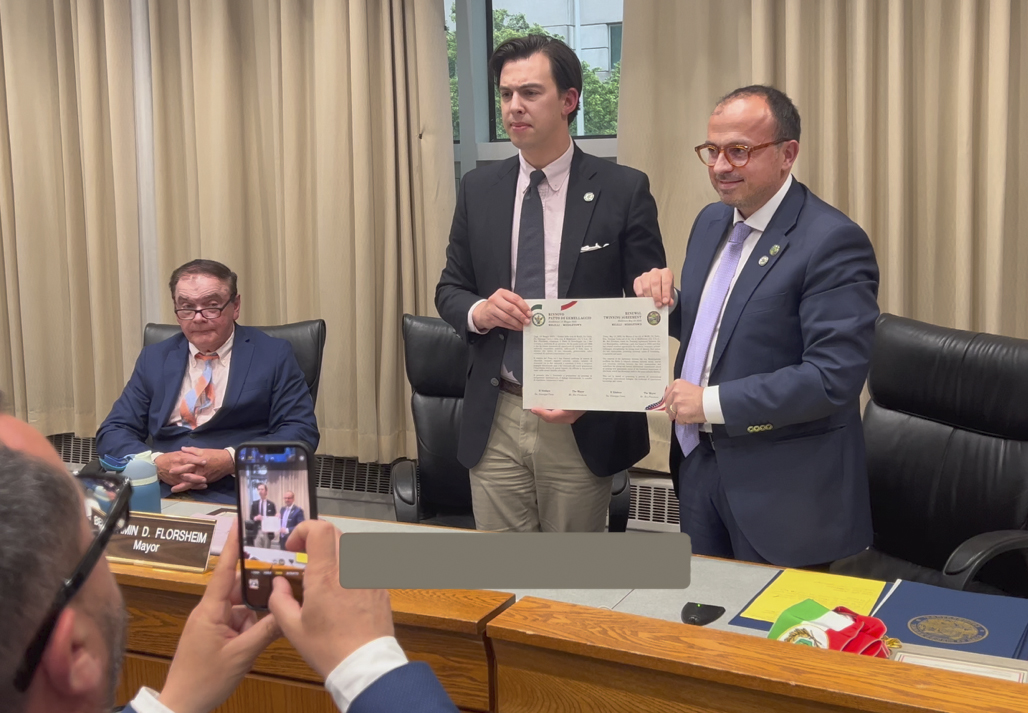 Two men in suits holding a framed document in a meeting room, while another person in the foreground takes a photo with a smartphone