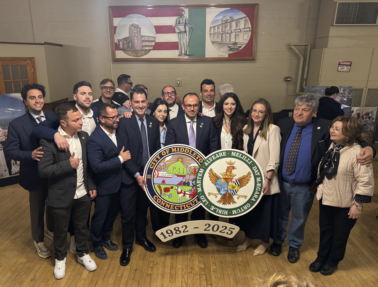 Group of people in formal attire holding a large emblem plaque reading ‘City of Middleboro, Connecticut — 1982-2025’ inside a hall”