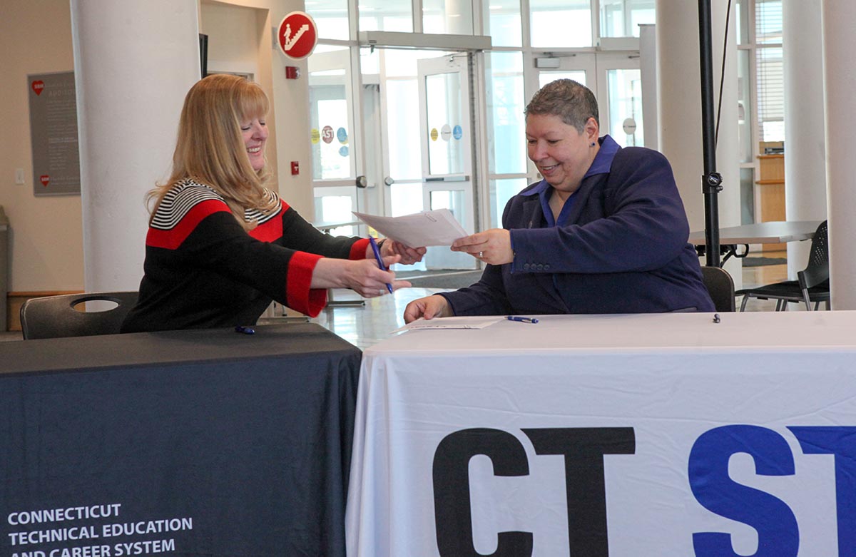 Photo by Brian Lombardo, CT State Manchester. President Christina Royal (right) smiling and handing a document across a table at a CT State and Connecticut Technical Education and Career System event.
