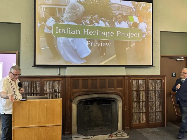 Two men present the "Italian Heritage Project Preview" in a room with a large projecOne man stands at a podium speaking into a microphone, while the other stands to the side holding a mic, near a fireplace.