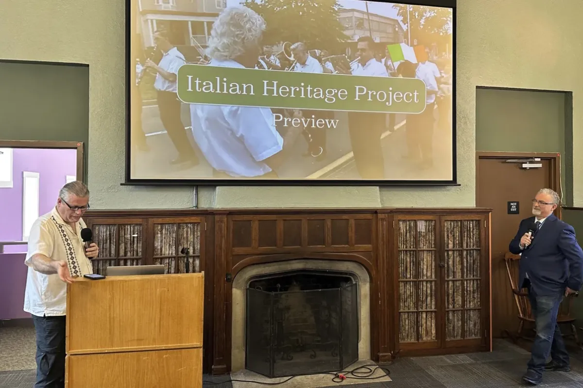 Two men present the "Italian Heritage Project Preview" in a room with a large projecOne man stands at a podium speaking into a microphone, while the other stands to the side holding a mic, near a fireplace.
