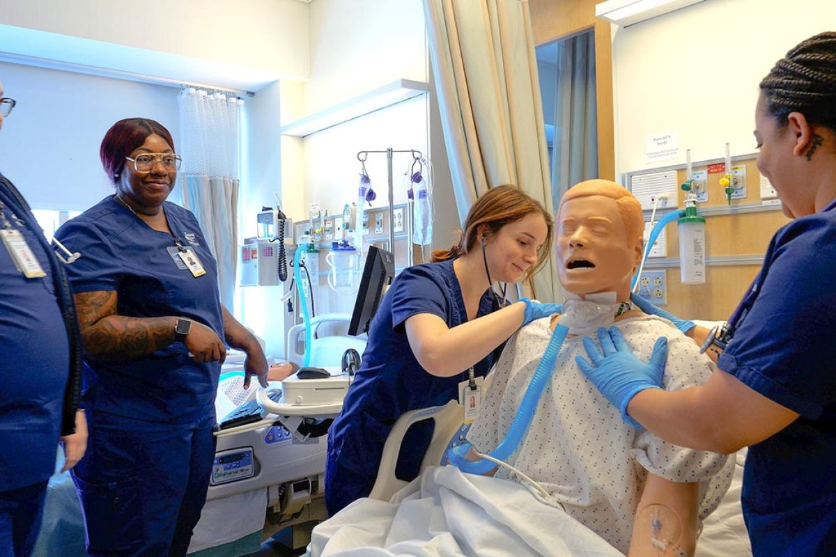 Nursing students in scrubs practicing CPR and airway management on a medical manikin in a clinical simulation room.