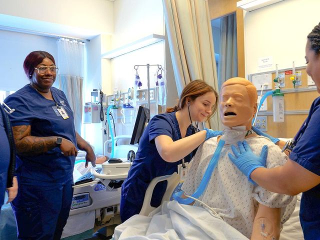 Nursing students in scrubs practicing CPR and airway management on a medical manikin in a clinical simulation room.