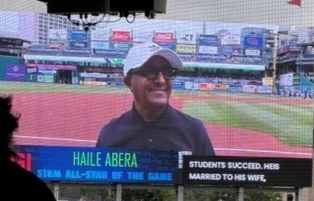 Man smiling in a baseball cap on big screen at a stadium