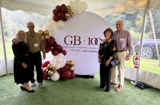 Four attendees pose beside a Goldblatt Bokoff 100th anniversary display decorated with burgundy, gold and white balloons.