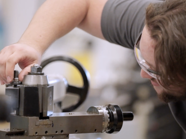 Student adjusting a metal lathe machine while wearing safety glasses in a workshop.