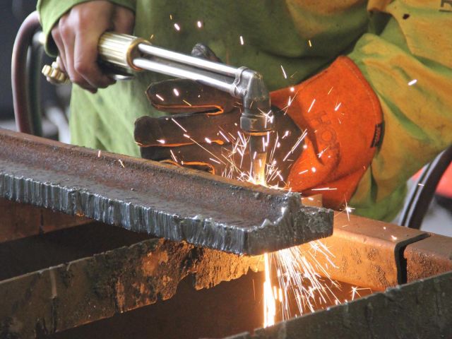 Worker wearing protective gloves using a torch to cut or weld a piece of metal, sparks flying in a manufacturing setting