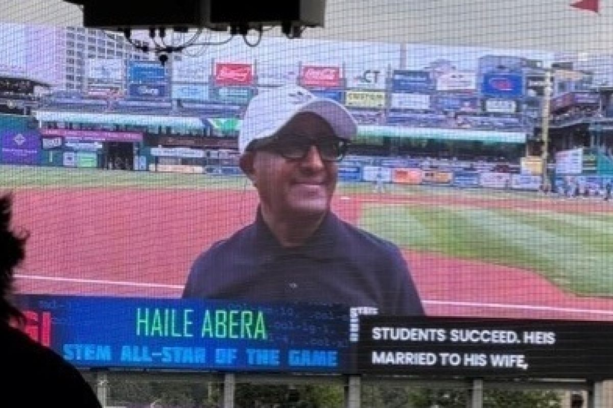 Man smiling in a baseball cap on big screen at a stadium