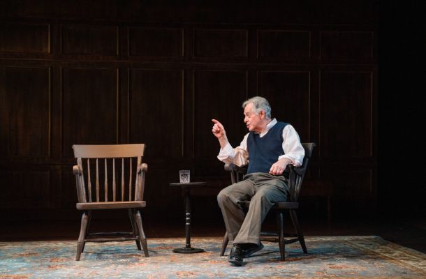 Man seated onstage in a wooden chair, gesturing toward an empty chair during a theatrical performance.