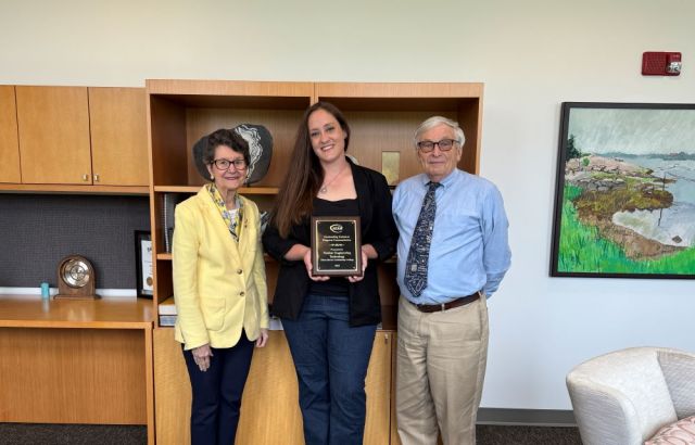 Three people stand together smiling. The middle person holds a plaque.